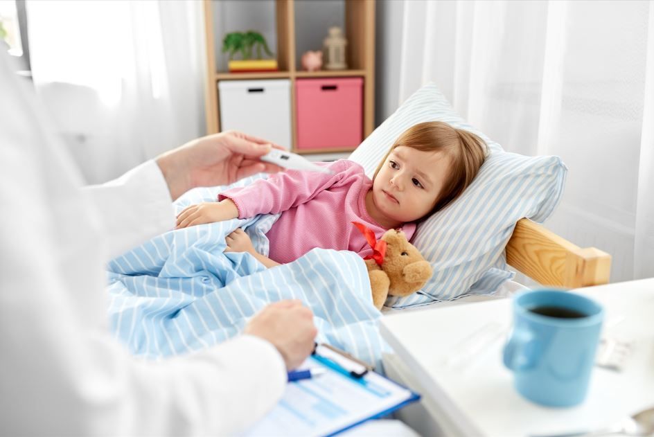 El médico le toma la temperatura al niño pequeño en la cama; el niño se ve enfermo, hay un osito de peluche cerca.