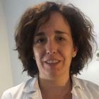 Una mujer con el pelo corto, castaño y rizado, que lleva una bata de laboratorio blanca, sonríe.