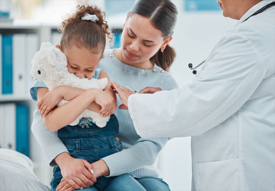 Niño abrazando un peluche, consolado por su madre durante un examen médico. Doctor con bata blanca.