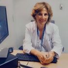 Una mujer con bata blanca está sentada en un escritorio con una computadora y un teclado, sonriendo.