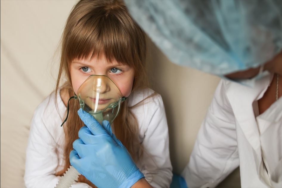 Niña con mascarilla de oxígeno, recibiendo asistencia de un profesional médico con guantes y gorro.