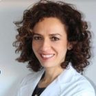 Mujer con cabello castaño rizado, vestida con un abrigo blanco, sonriendo.