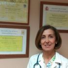 Mujer con bata blanca y estetoscopio, sonriendo, delante de unos certificados enmarcados.