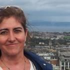 Mujer sonríe, fondo urbano con cielo nublado, lleva una chaqueta azul y gafas de sol en la cabeza.