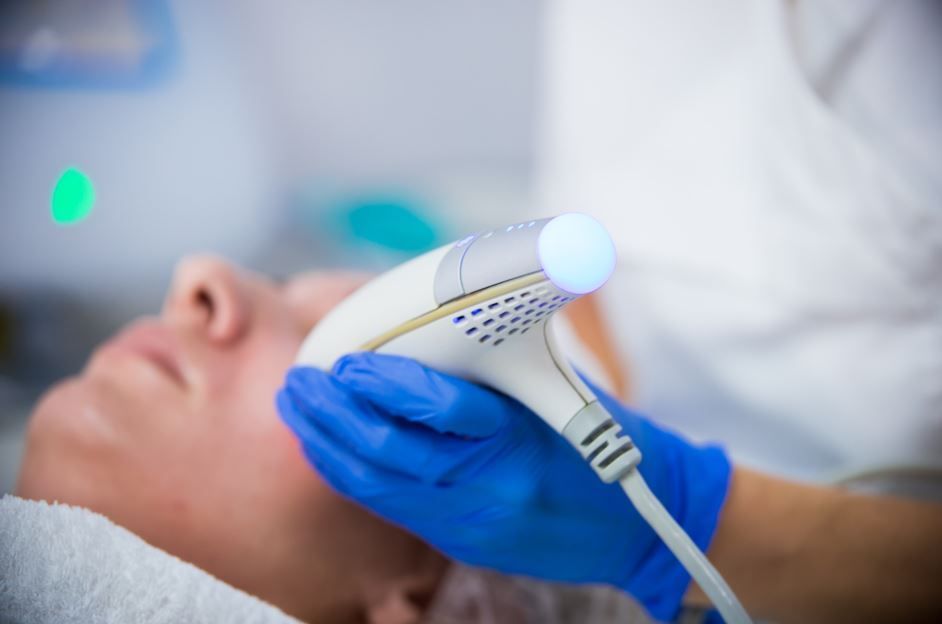 Persona recibiendo tratamiento facial con un dispositivo manual, guantes azules, toalla blanca, luces brillantes.