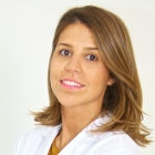 Mujer con cabello castaño hasta los hombros, sonriendo, vestida con un abrigo blanco, mirando a la cámara.