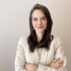 Mujer con los brazos cruzados, vestida con una chaqueta de color claro, sonriendo, sobre un fondo liso.