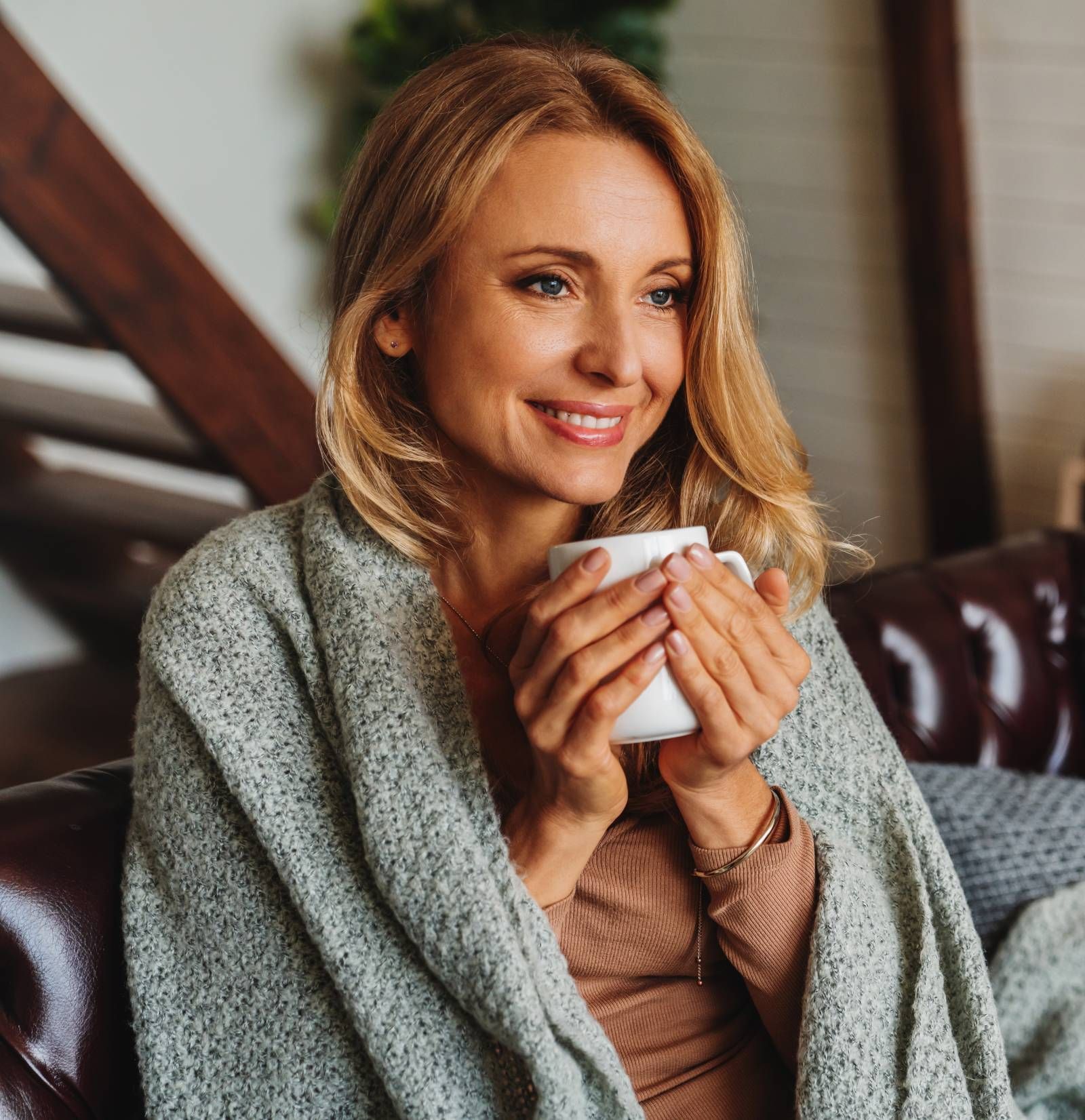 Frau, in eine graue Decke gehüllt, lächelnd, mit einer weißen Tasse auf einem Ledersofa.