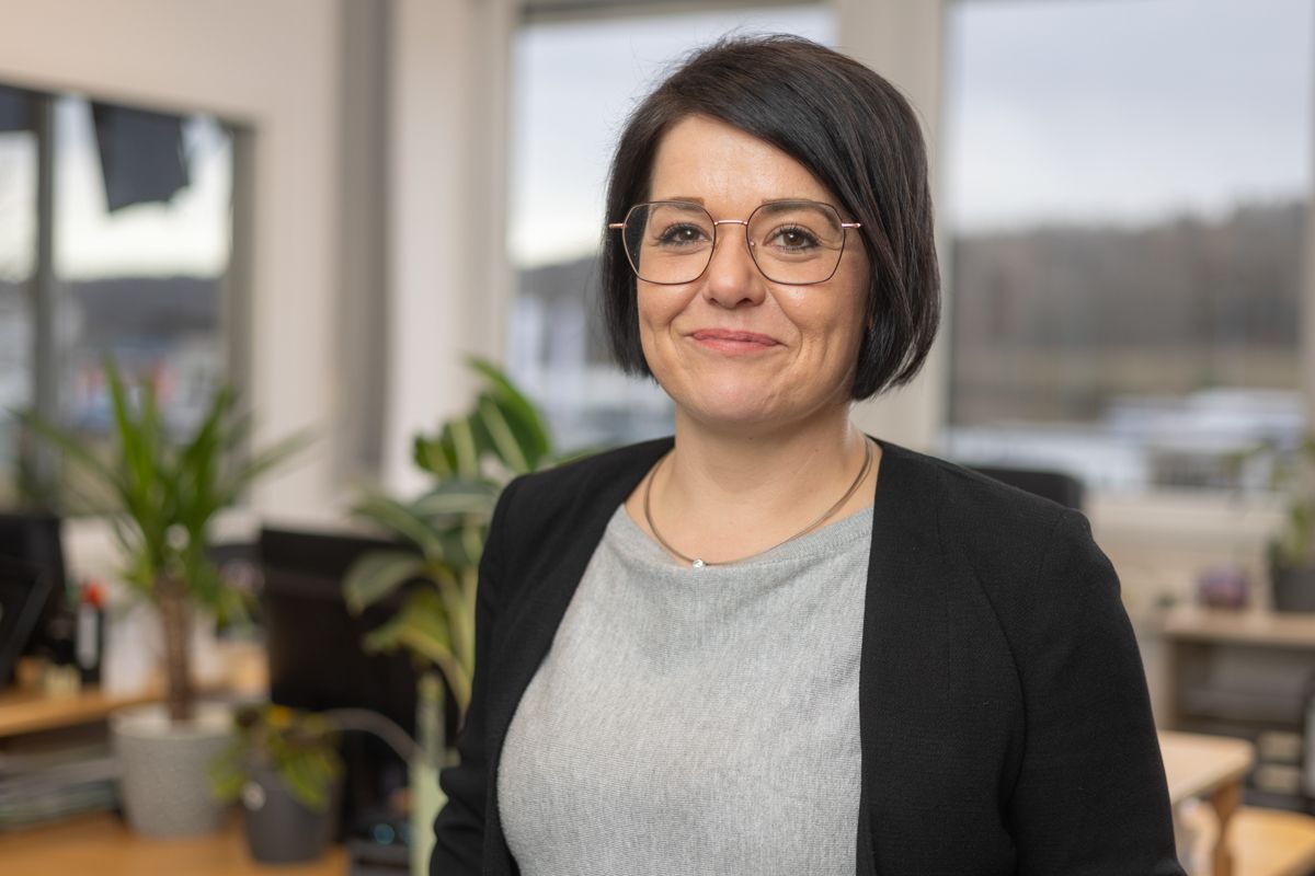 Eine Frau mit Brille und schwarzer Jacke lächelt in einem Büro.