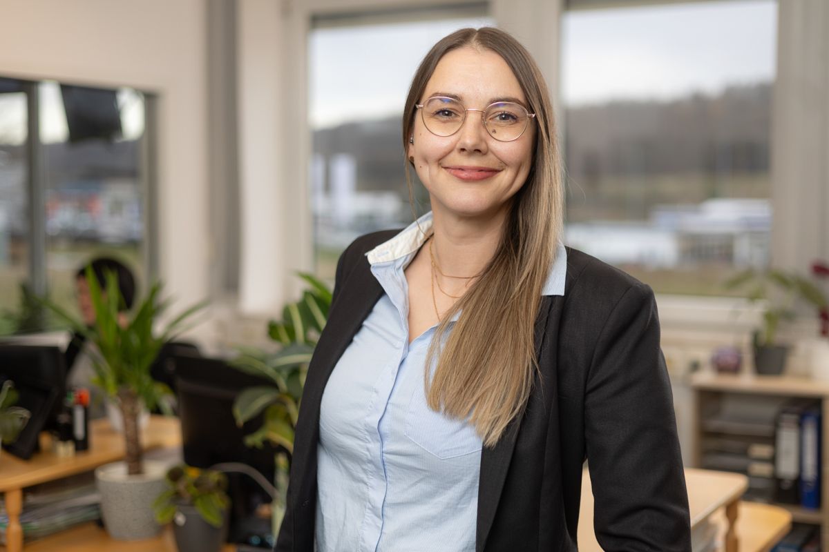 Eine Frau in Anzug und Brille steht vor einem Fenster in einem Büro.