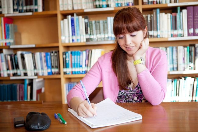 Mujer con un cárdigan rosa escribiendo en un escritorio de biblioteca, rodeada de estanterías.