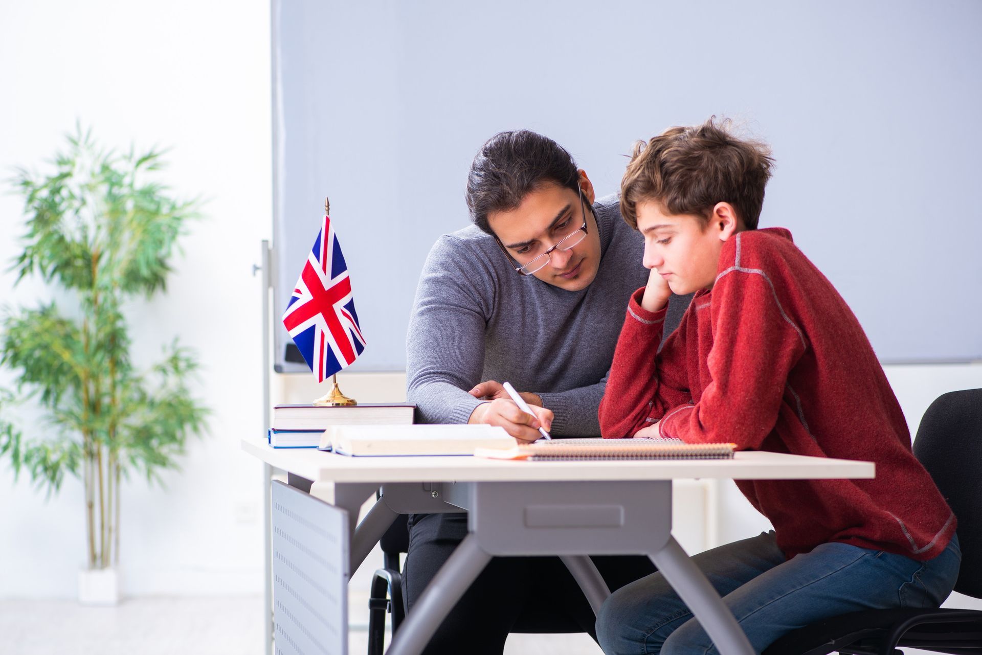 El tutor ayuda a un estudiante con la tarea en el escritorio; la bandera británica está sobre la mesa.