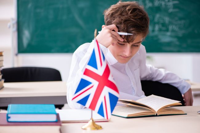 Un niño en un aula con una bandera del Reino Unido estudia, sostiene un bolígrafo y mira un libro cerca de una pizarra.