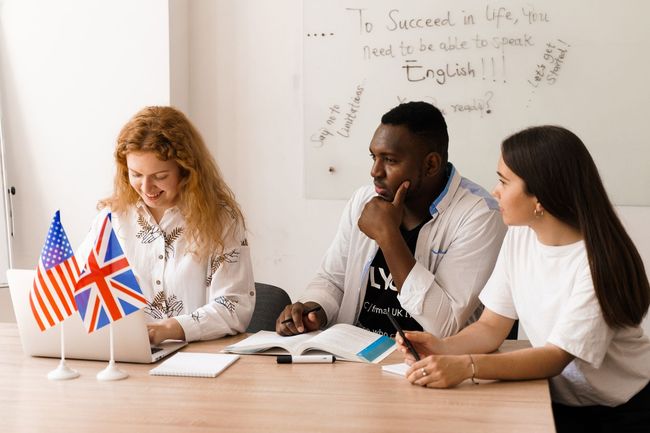 Tres personas en una clase de inglés, mirando una computadora portátil y un libro abierto.