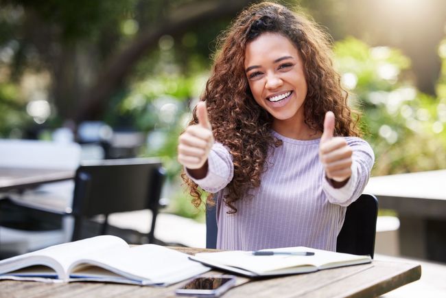 Mujer joven sonriente dando dos pulgares hacia arriba, estudiando al aire libre.