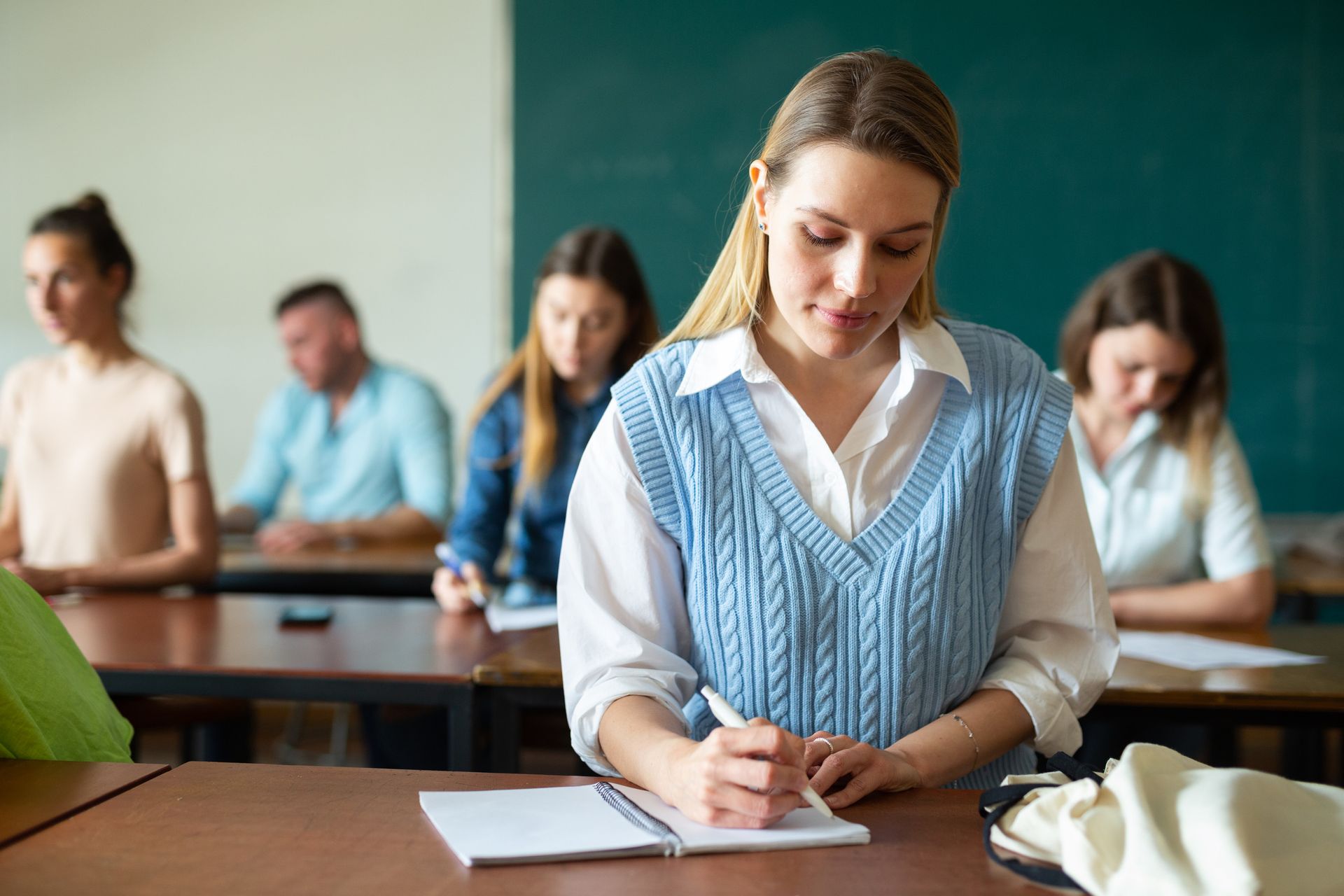 Mujer con un chaleco azul escribiendo en un cuaderno en un escritorio en un aula, otros estudiantes detrás de ella.