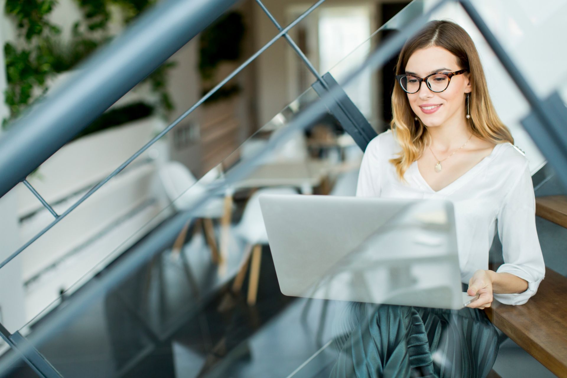Una mujer con gafas trabaja en una computadora portátil mientras está sentada en las escaleras de una oficina moderna.