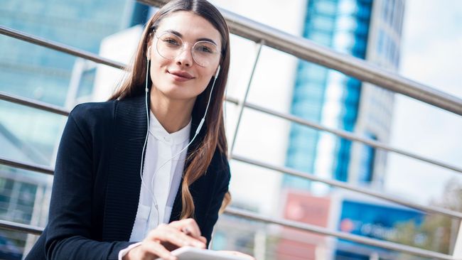 Mujer vestida de negocios, con gafas y auriculares sonríe a la cámara al aire libre con una ciudad de fondo.