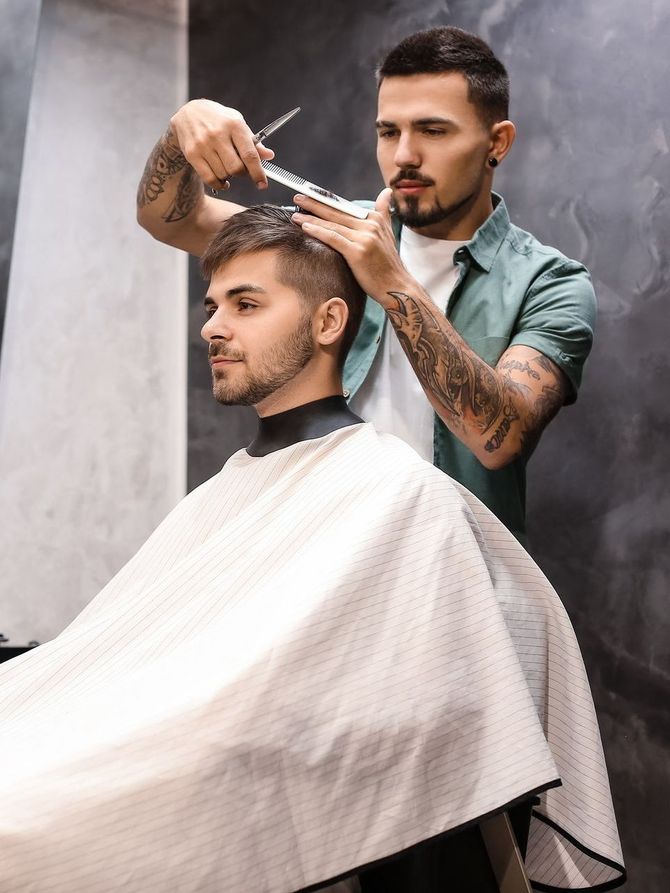 Un joven sonriente con el pelo engominado hacia atrás y una camisa gris. Al fondo, un hombre con camisa gris. Interior.