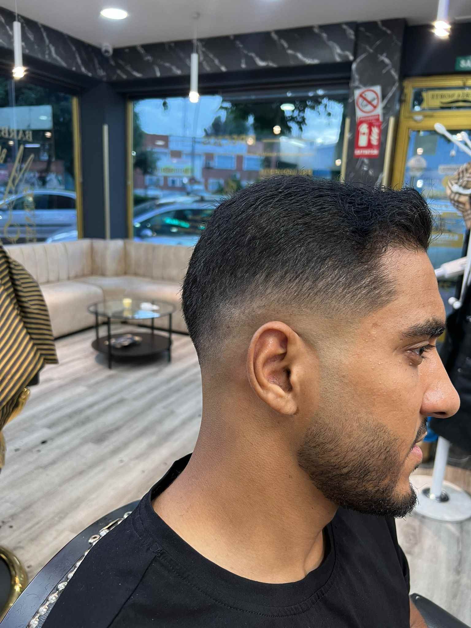 Un hombre con un corte de pelo degradado en una barbería. Lleva una camisa negra, y la tienda tiene suelo de madera clara y una pared oscura.