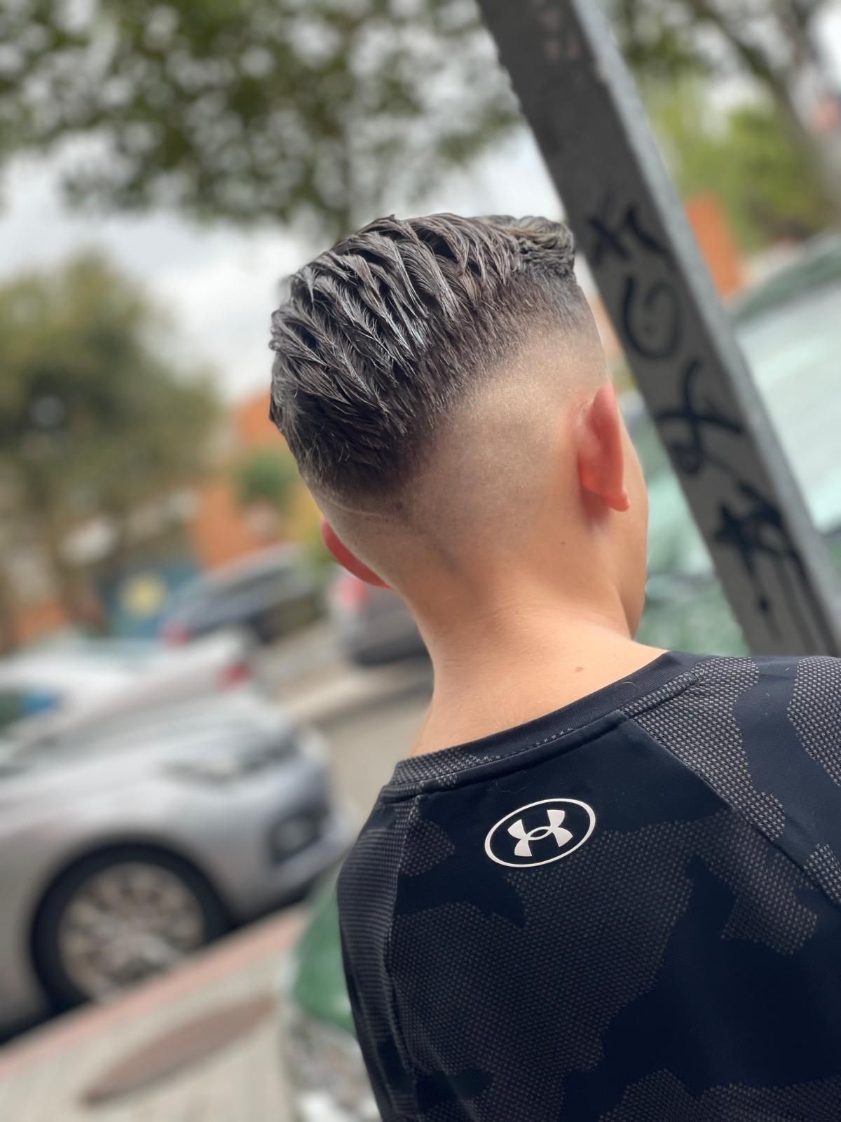 Persona joven con corte de pelo, de espaldas a la cámara, vistiendo una camiseta negra de Under Armour, al aire libre con coches y grafitis visibles.