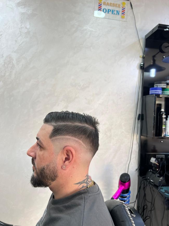 Hombre con barba y corte de pelo descolorido en una barbería. Lleva la raya al lado y un diseño rapado.