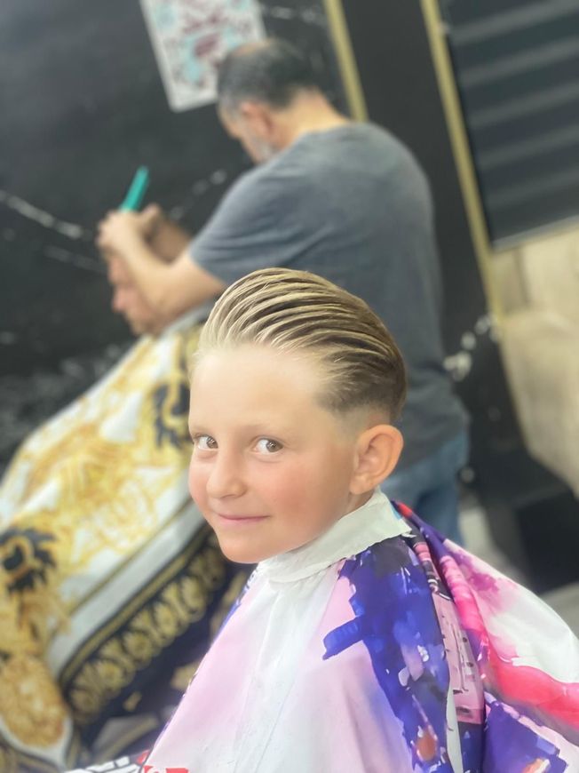 Un joven con el pelo engominado hacia atrás sonríe a la cámara, con una capa de barbero. Al fondo, otra persona corta el pelo.