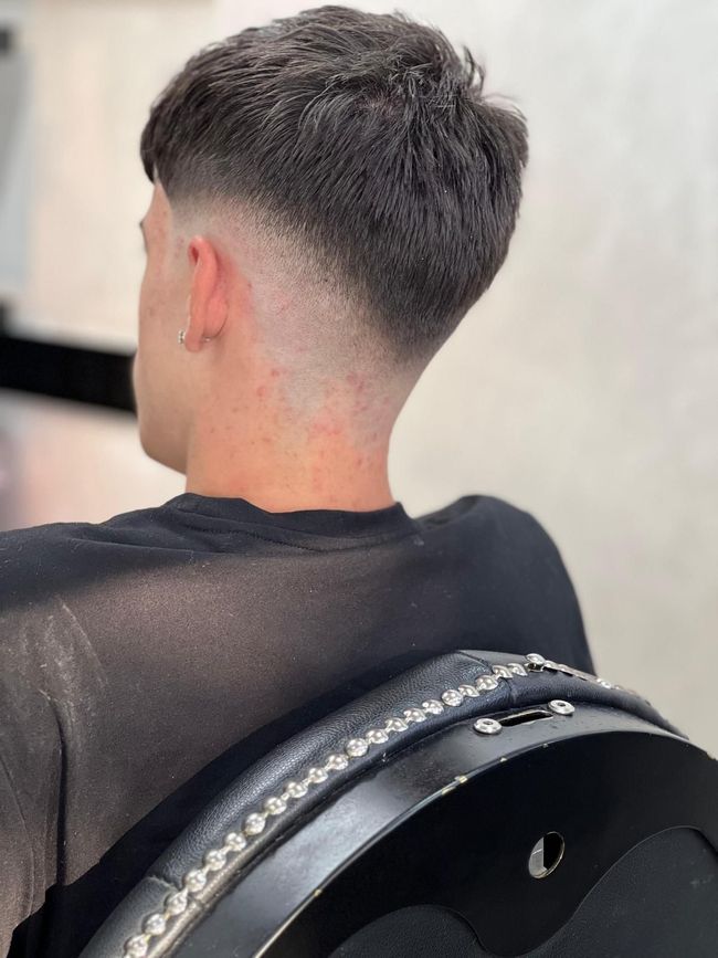 Hombre con corte de pelo corto y descolorido, cabello oscuro en la parte superior y desvanecido claro en los costados y la espalda, sentado en una silla.
