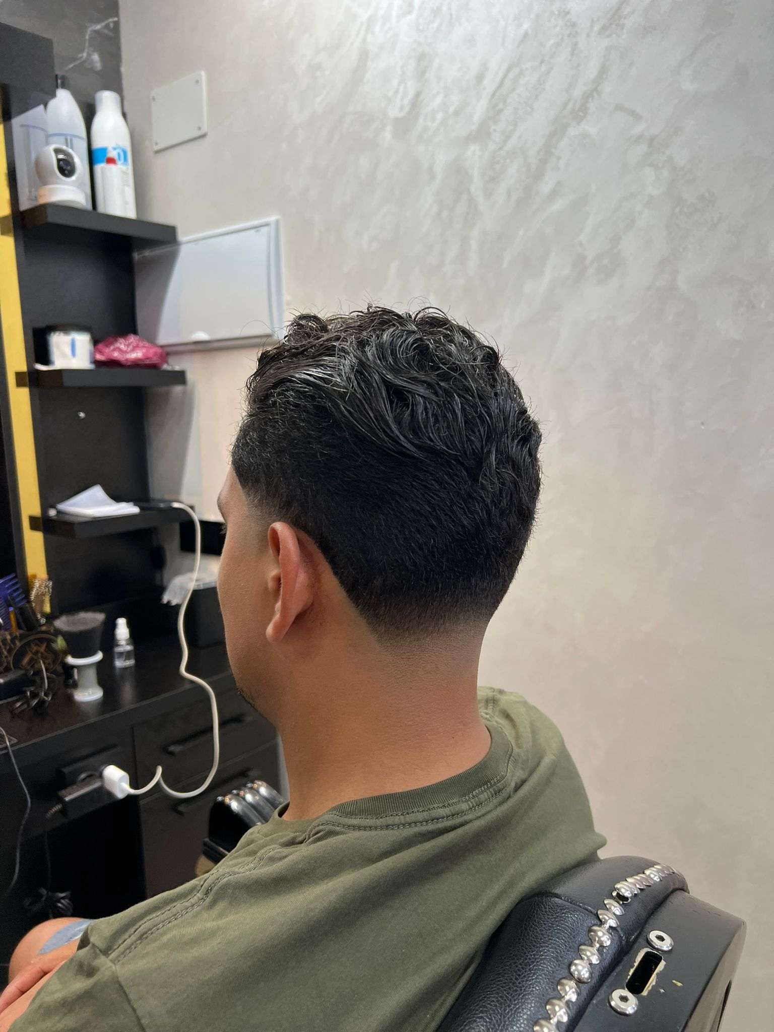 Hombre cortándose el pelo en una barbería. Primer plano de la nuca. Lleva el pelo oscuro y bien peinado, y lleva una camisa verde.