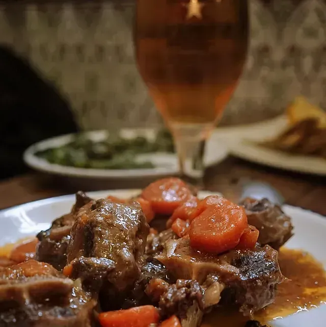 Un plato de carne y zanahorias con un vaso de cerveza al fondo.