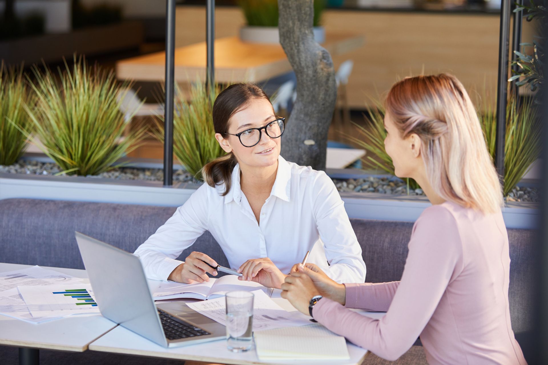 Dos mujeres en una mesa al aire libre; una con camisa blanca y gafas, gesticulando; la otra de rosa, cogidas de la mano. Ordenador portátil y papeles.