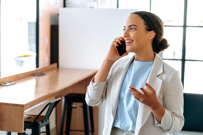 Una mujer con chaqueta habla por teléfono sonriendo, apoyada en una encimera de madera cerca de la ventana.