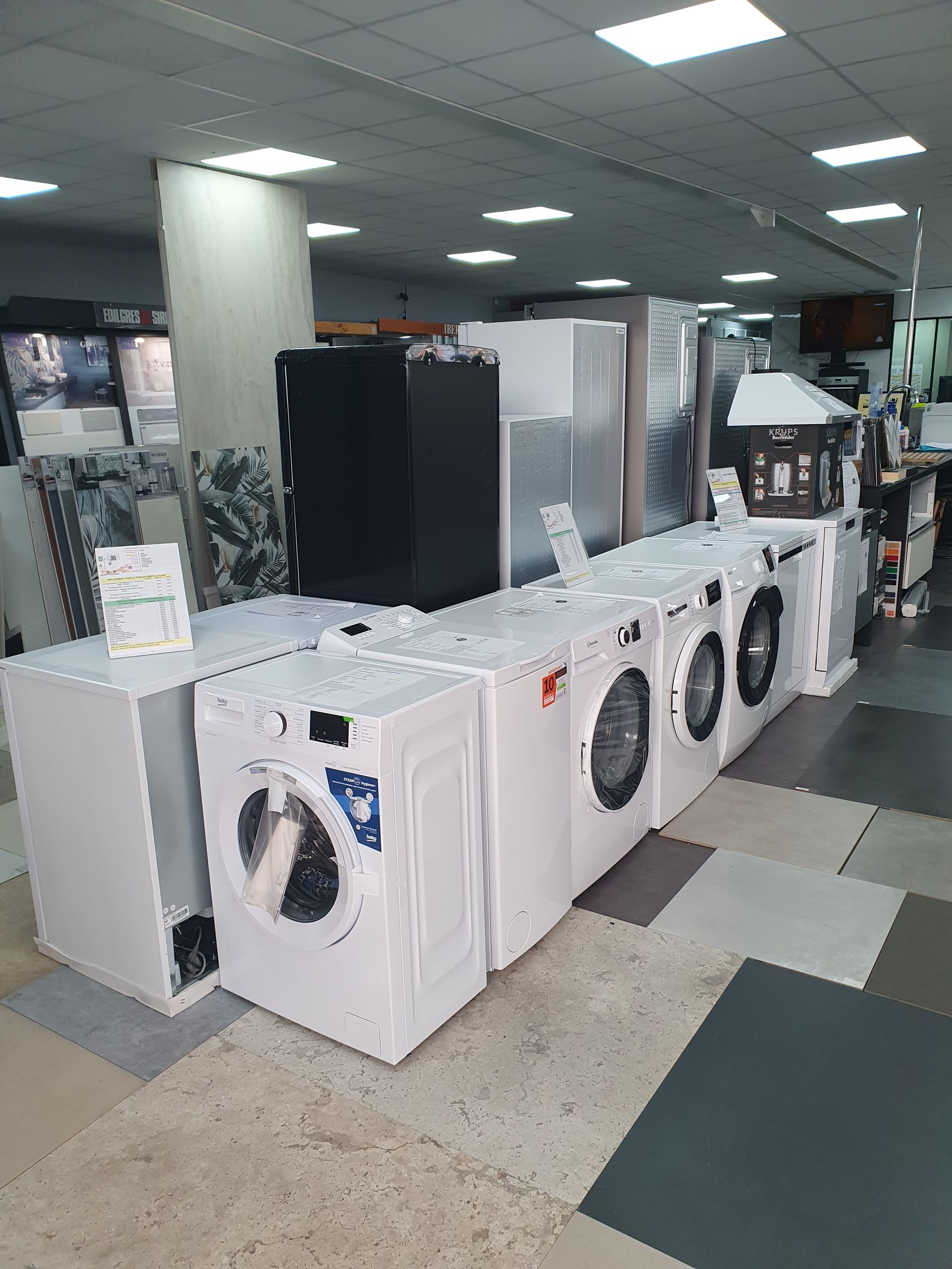 Machines à laver et réfrigérateurs exposés dans un magasin d'électroménager. Des appareils blancs sont alignés, d'autres appareils étant à l'arrière-plan.