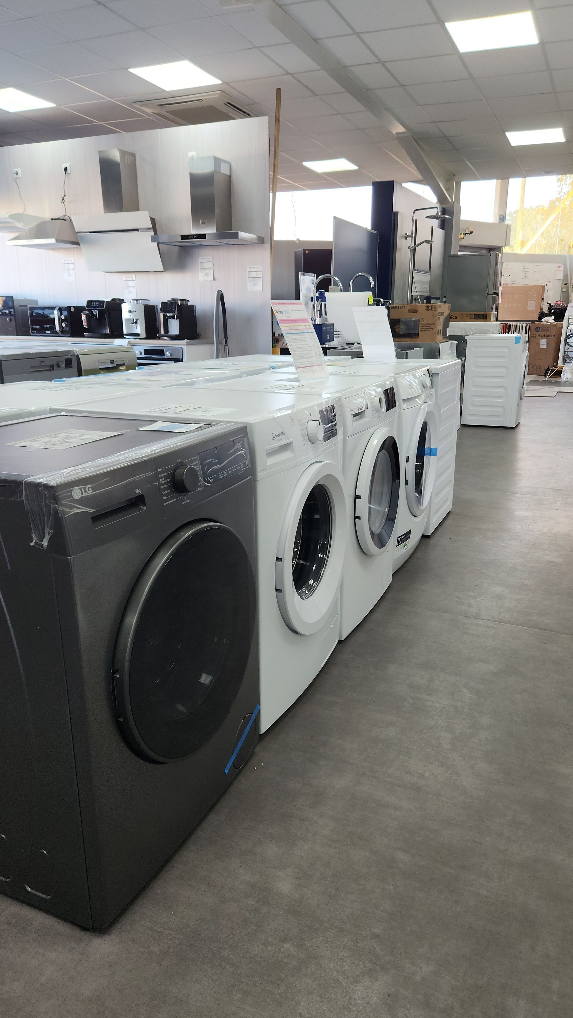 Rangée de machines à laver dans un magasin d'électroménager très éclairé. Les machines sont de couleurs variées