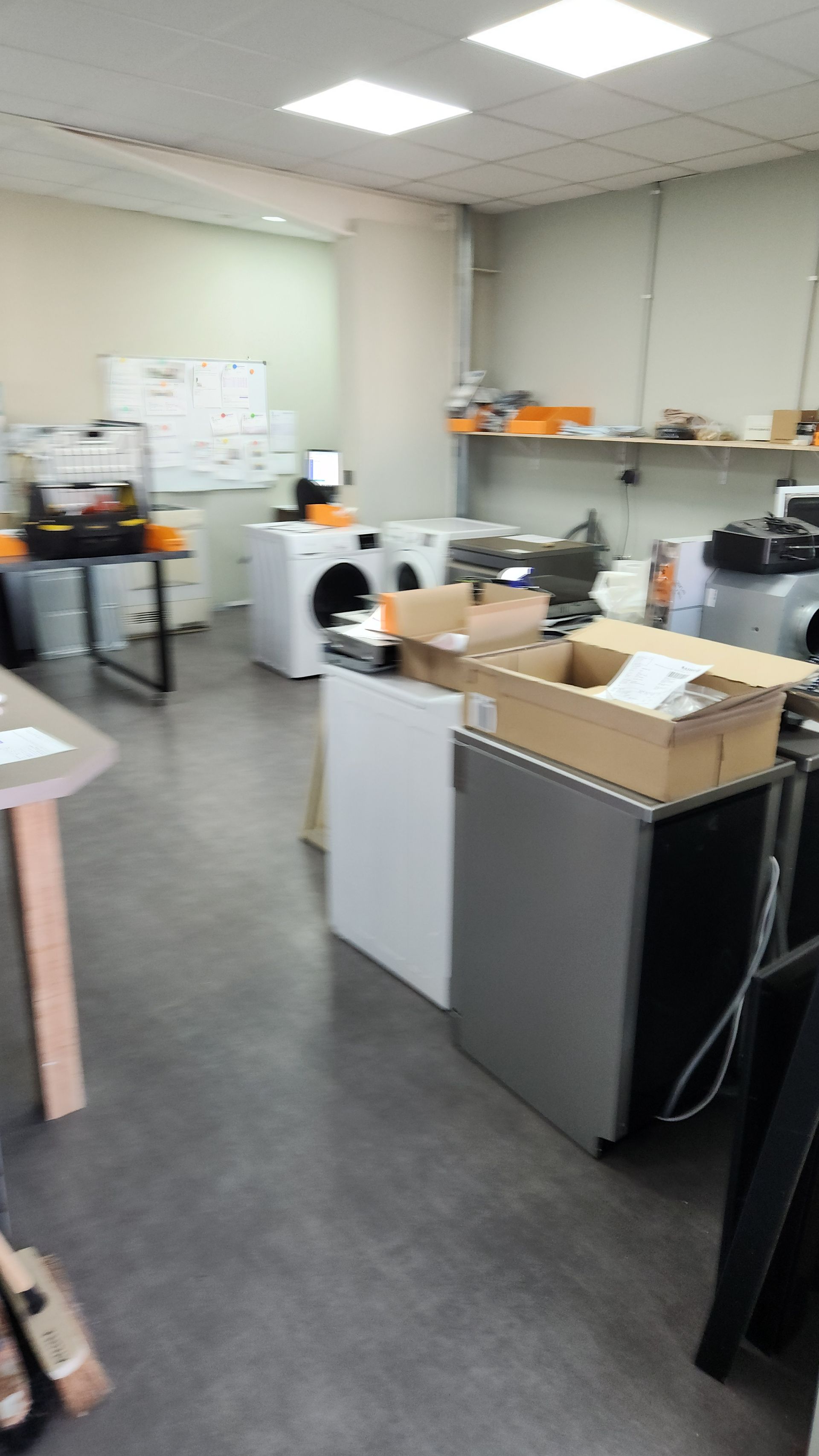 Un atelier de réparation proposant divers appareils électroménagers, tels que des machines à laver et des réfrigérateurs. Des cartons et un tableau blanc sont également présents.