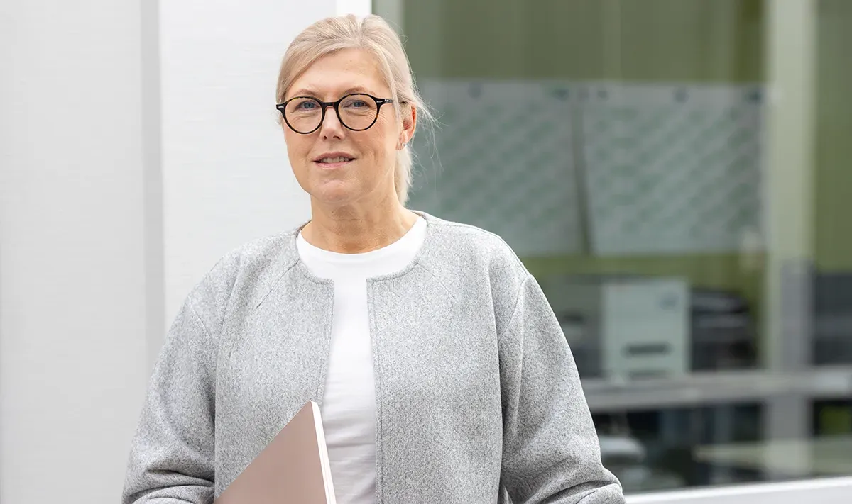 Frau mit Brille und grauer Jacke, die einen Laptop hält, steht draußen am Fenster.