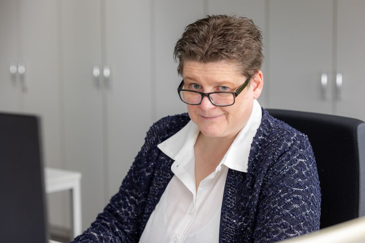 Frau mit Brille, die an einem Schreibtisch sitzt, in die Kamera schaut und einen Blazer über einem weißen Hemd trägt.