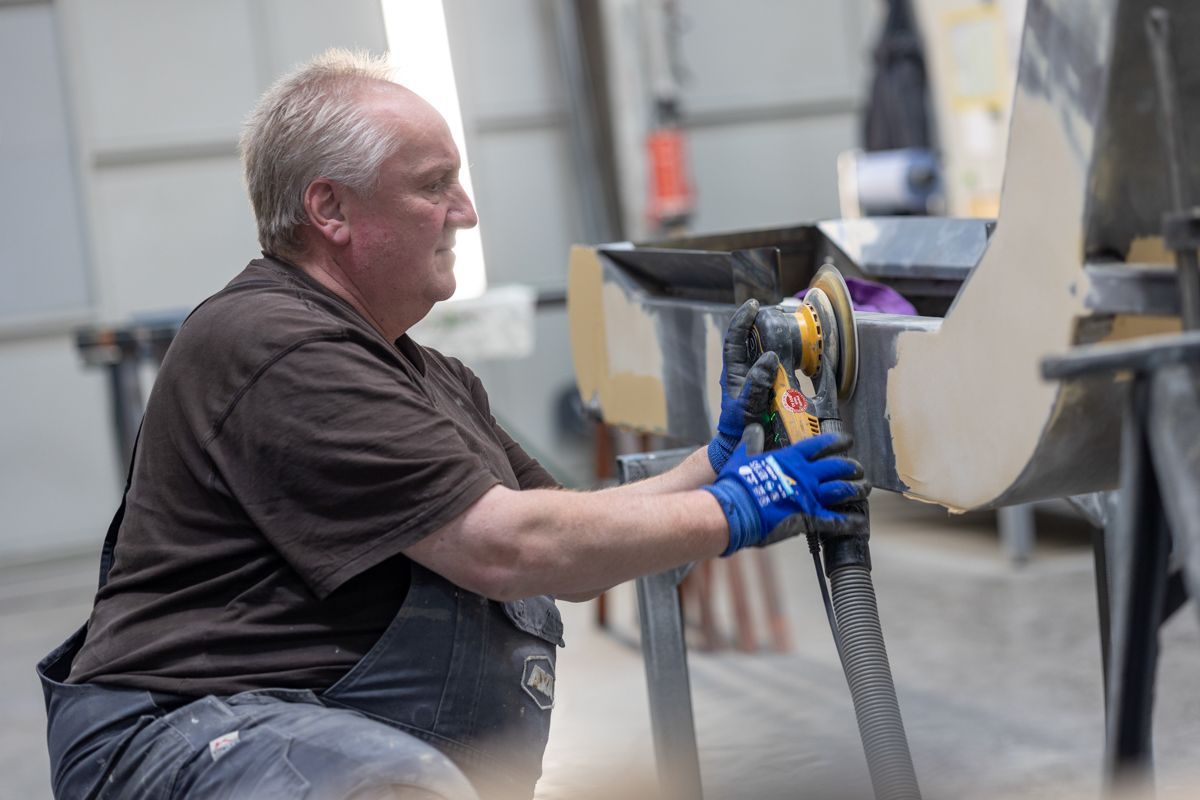 Ein Mann in einer Werkstatt, der mit einem Schleifgerät an einem gebogenen Gegenstand arbeitet, trägt Arbeitshandschuhe.