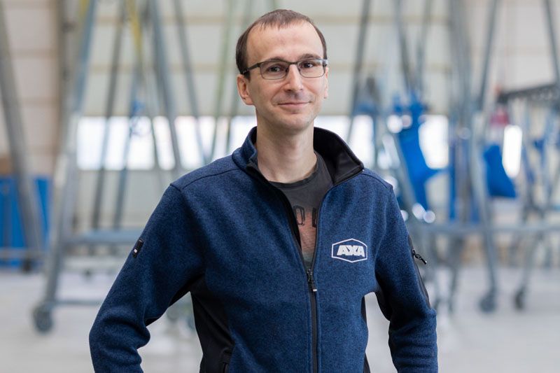 Ein Mann mit Brille lächelt in einer Fabrikhalle. Er trägt eine blaue Jacke mit Logo.