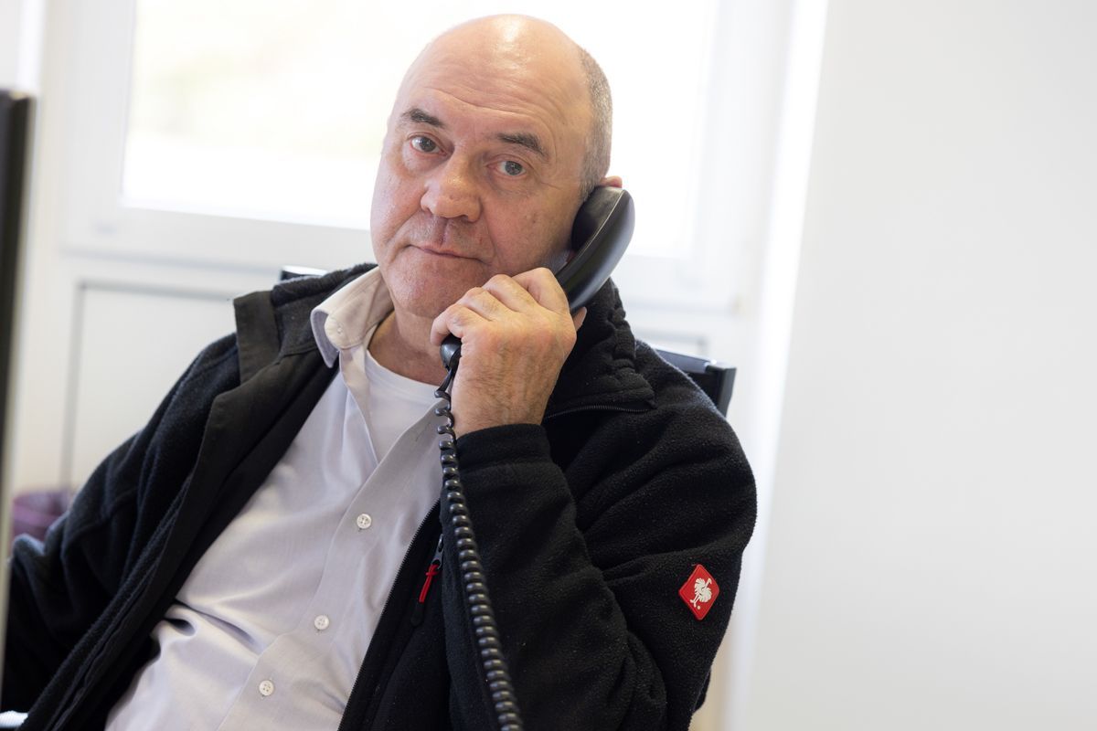 Ein Mann, der in einem Büro ein Festnetztelefon hält, in die Kamera blickt, trägt eine schwarze Jacke und ein weißes Hemd.