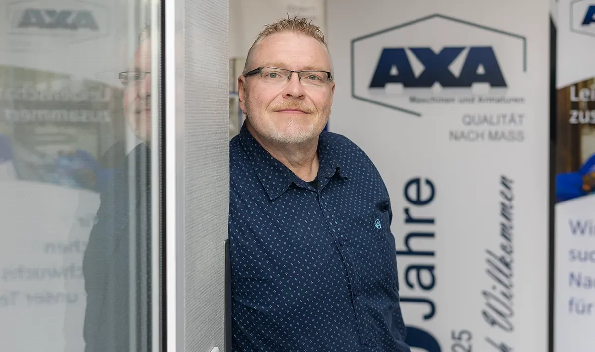 Mann in blauem Hemd und Brille, lächelnd, steht neben einem AXA-Banner.