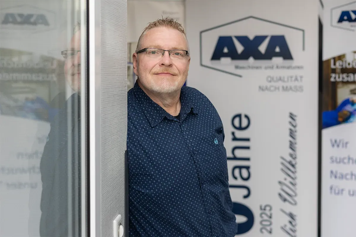Ein lächelnder Mann in blauem Hemd steht neben einem „AXA“-Schild, möglicherweise am Eingang eines Geschäfts.