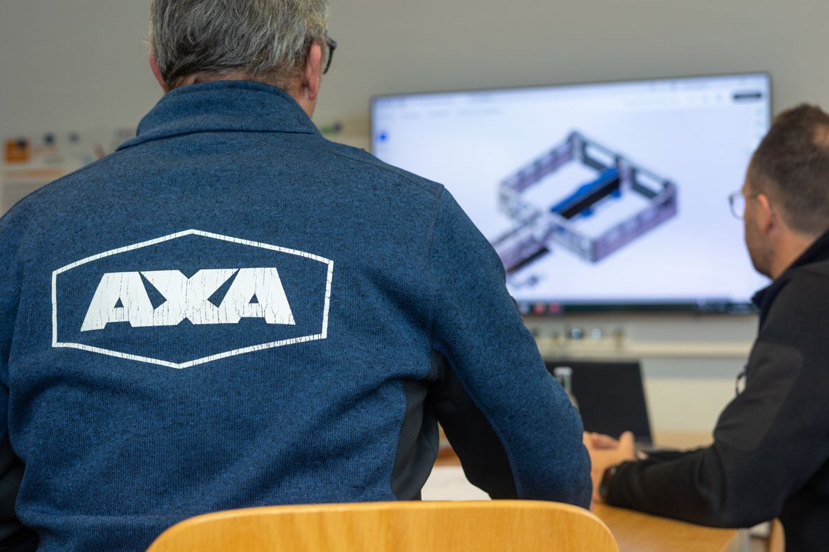 Zwei Männer an einem Computer, einer davon trägt eine Jacke mit AXA-Logo, begutachten eine 3D-Konstruktion eines mechanischen Bauteils.