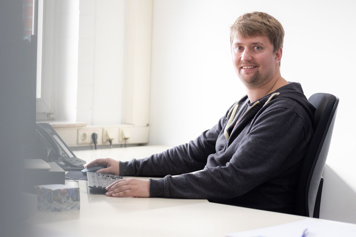 Ein Mann in grauem Kapuzenpulli sitzt lächelnd an einem Schreibtisch. Hinter ihm befinden sich eine weiße Wand und ein Fenster, auf dem Schreibtisch stehen ein Computer und eine Tasse.