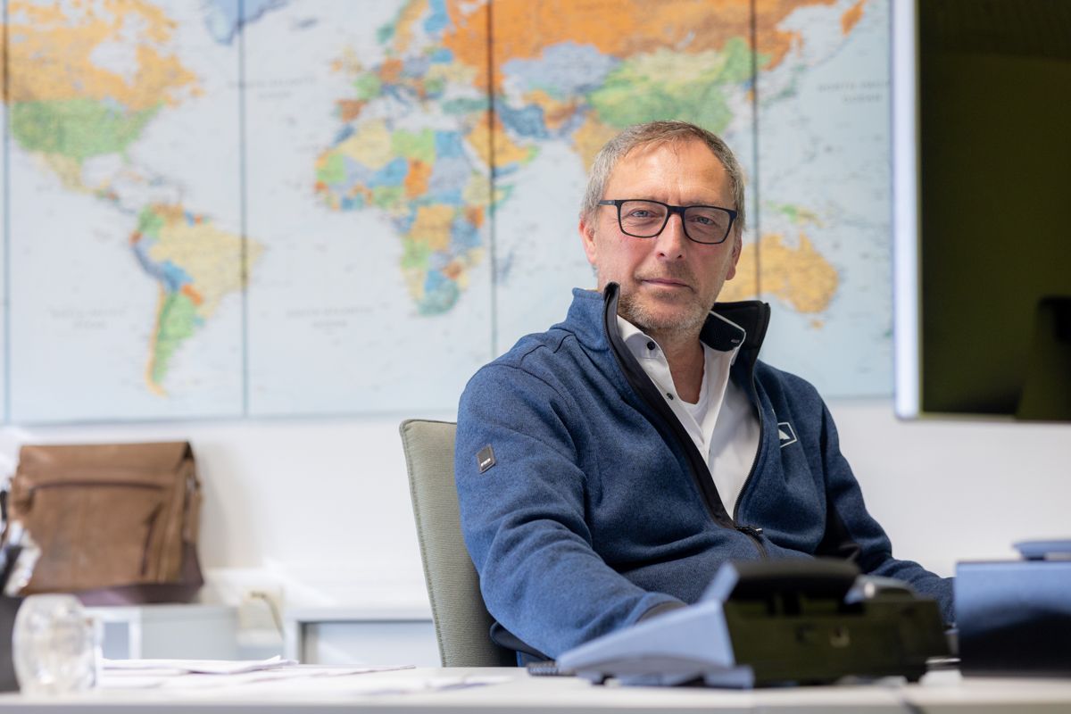 Mann mit Brille sitzt an einem Schreibtisch und blickt in die Kamera. Hintergrund: Weltkarte.