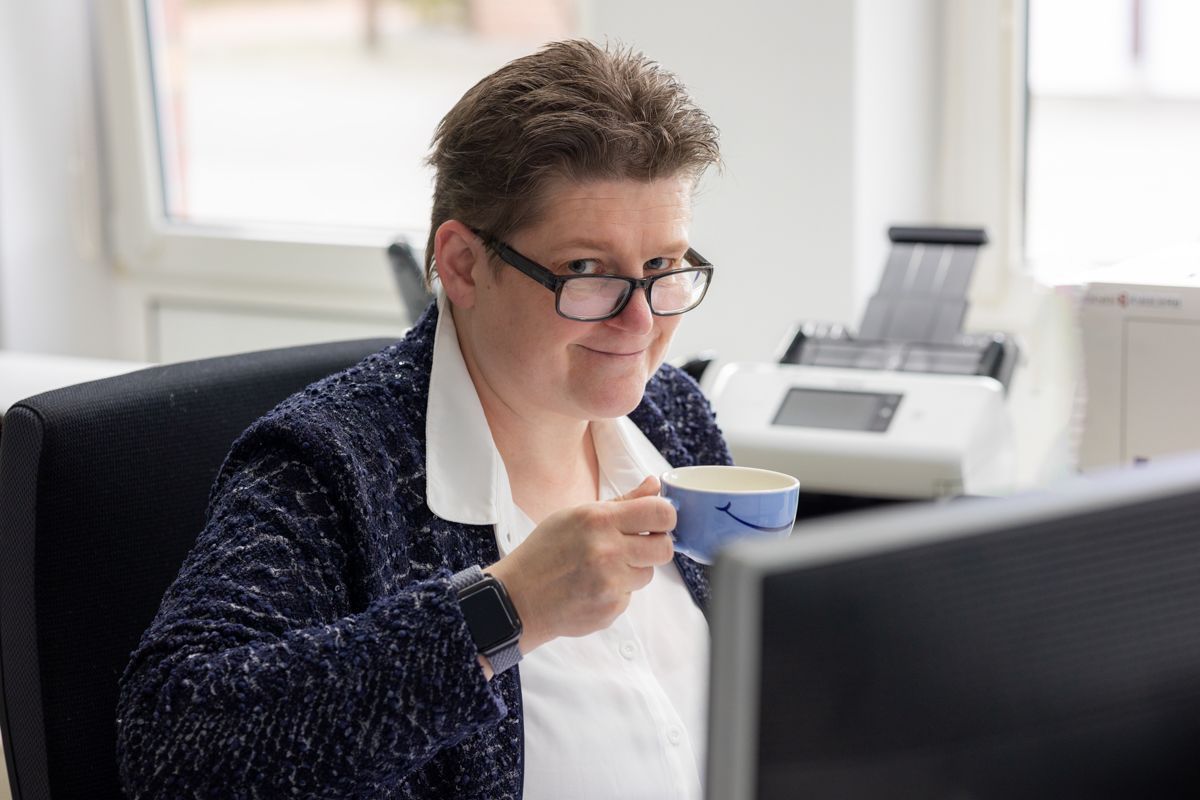 Eine Frau mit Brille, die eine Kaffeetasse hält, lächelt in die Kamera, während sie an einem Schreibtisch mit Computer und Drucker sitzt.