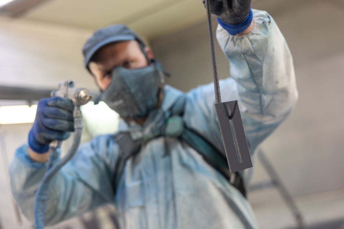 Eine Person in Schutzkleidung besprüht einen dunklen Gegenstand mit Farbe; der Hintergrund ist verschwommen.