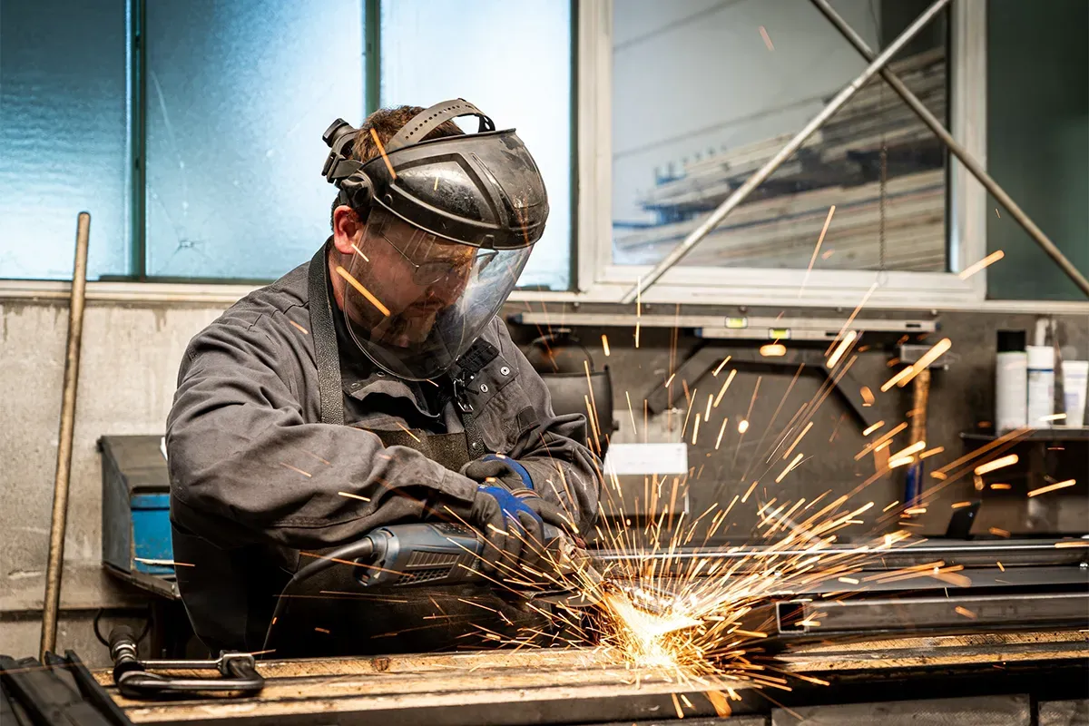 Ein Schweißer in einer Werkstatt schleift Metall, Funken sprühen, er trägt Schutzausrüstung.
