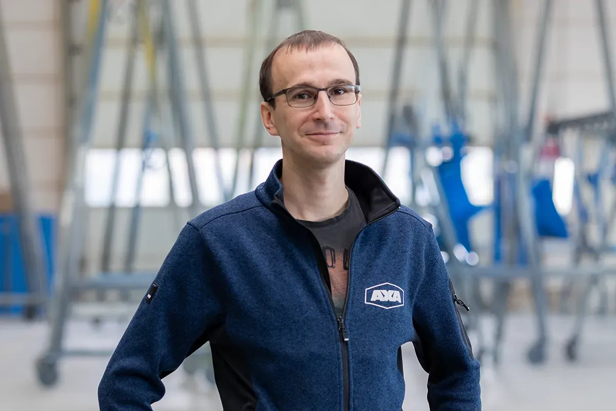 Ein Mann mit Brille und blauer Jacke lächelt in einer Fabrikhalle.