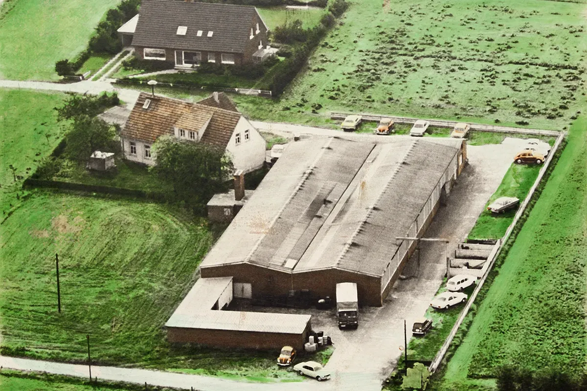 Luftaufnahme eines Industriegebäudes und zweier Häuser, umgeben von Feldern und Straßen. Draußen parken Autos.